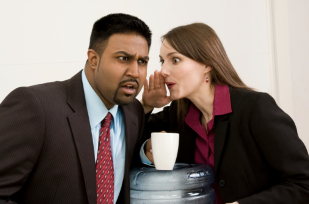 a women in business atire whispering to her male coworker at the water cooler.