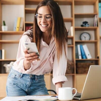 An individual looking at their phone while leaning on the table with a laptop. the image is used to indicate someone using AI to build smarter meeting agenda's.
